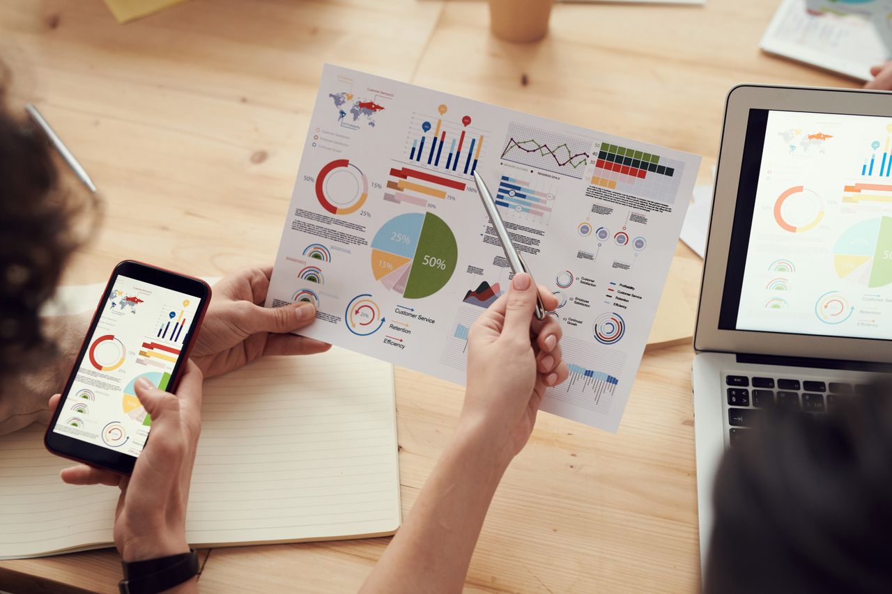 Professionals reviewing financial charts and dashboards across paper, phone and laptop.